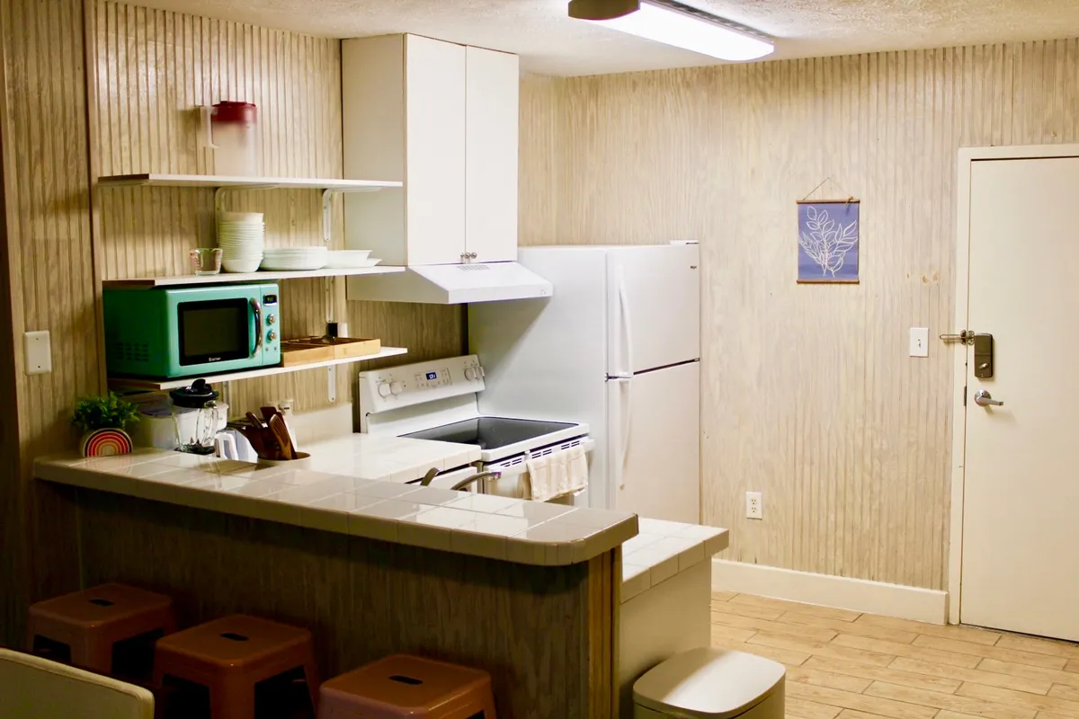 A cozy kitchenette space featuring a compact kitchen island, a visible microwave and refrigerator, and open shelves stocked with essential dishware. The scene suggests meal preparation with a casual, inviting ambiance, enhanced by natural light and homely touches.