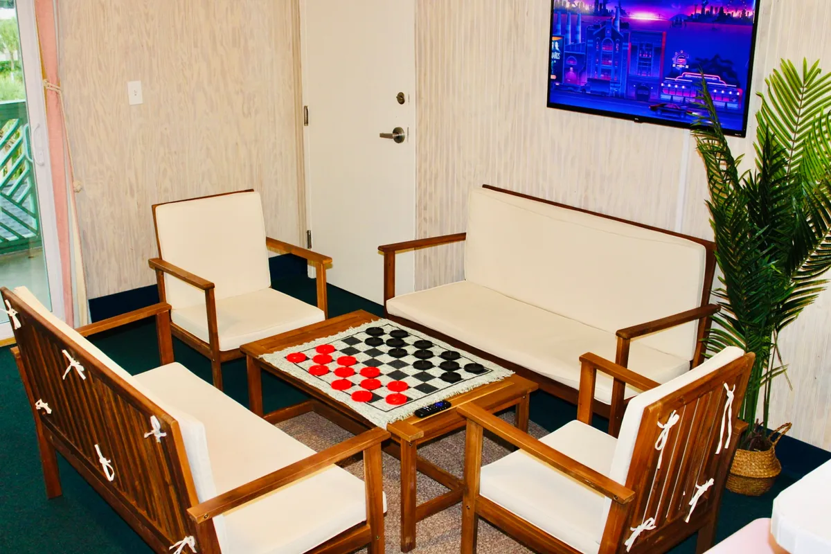 This image shows a welcoming interior space featuring a checkerboard table with comfortable seating, a television, and a calming indoor plant. The room radiates a friendly, relaxed mood, making it an inviting space for guests to unwind, socialize, or enjoy a cozy night in.