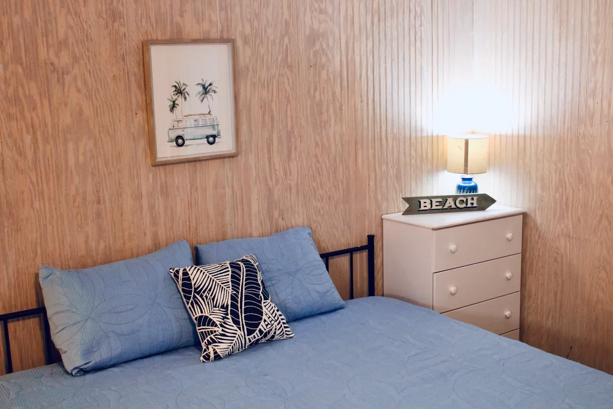 Featuring a bed with blue bedding, a decorative pillow, a lamp with a beach sign, and framed artwork, this tranquil scene provides a glimpse of the studio's restful ambiance. The decor leans into a beachside theme, enhancing the sense of location and relaxation.