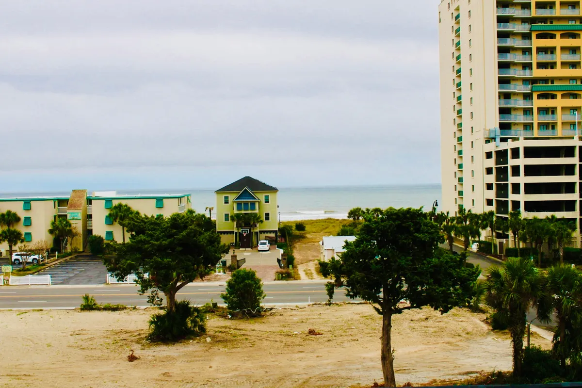 This image features a tranquil ocean view with a green building in the foreground, framed by tall buildings and trees. The scene suggests proximity to the shoreline and offers a glimpse of the relaxing beach environment just steps away.