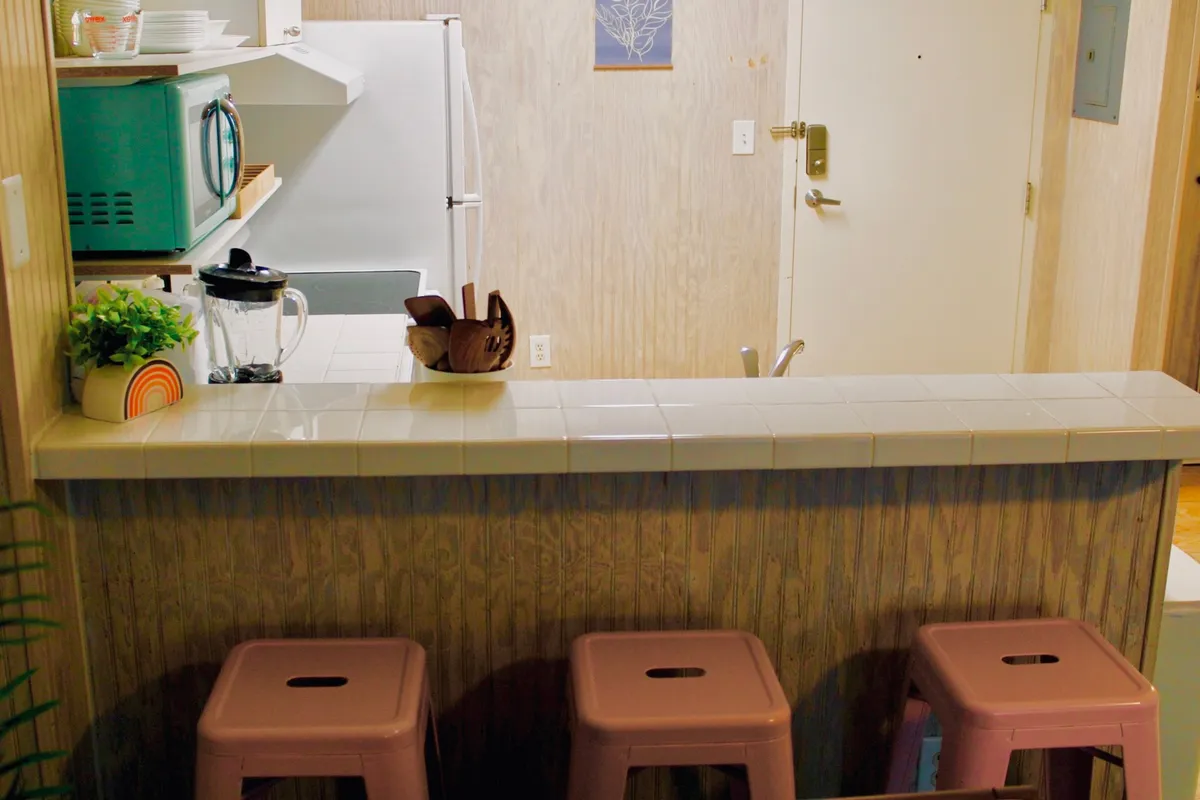 This cheerful interior scene features a kitchen island with colorful stools, a blender, and vibrant wall artwork. The setup is perfect for casual dining, socializing, or whipping up a quick meal. The open layout allows the dining and kitchen areas to seamlessly merge, aligning with the studio efficiency concept.